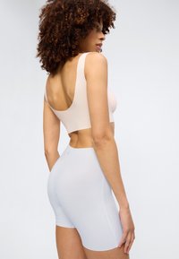 Femme aux cheveux bouclés portant un haut beige sans manches et un short ajusté bleu clair, debout de profil sur un fond uni.