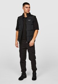 Zwarte puffer vest met een geborduurd ontwerp, hoge kraag en rits aan de voorkant. Draag het over een zwart T-shirt met korte mouwen en in combinatie met zwarte cargobroeken.