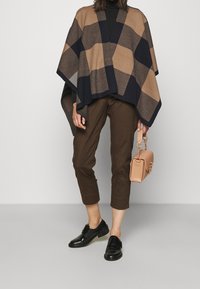 Brun och marinblå rutig poncho över en svart polotröja, kombinerad med bruna croppade byxor och svarta skor. Håller en liten beige handväska.