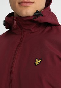 Bordowa kurtka z kapturem, z zamkiem z przodu, regulowanym kapturem ze sznurkiem oraz małym żółtym haftowanym logo na piersi. Gładka faktura materiału.