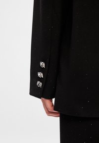 Mâneca unui sacou negru cu nasturi metalici și embellismente împrăștiate pe un material texturat. Prim-plan al detaliilor îmbrăcămintei.