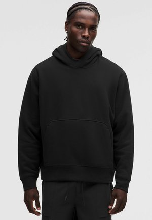 Mann mit geflochtenem Haar, der einen schlichten schwarzen Kapuzenpullover und passende schwarze Hose vor neutralem Hintergrund trägt.