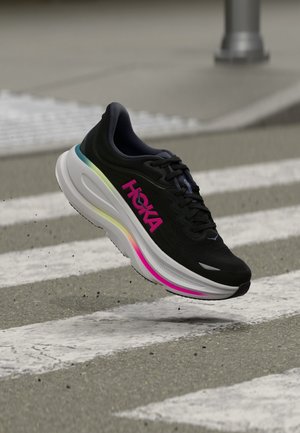Black running shoe with pink HOKA logo and multicolored sole mid-air above a crosswalk on a paved street.