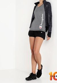 Conjunto atlético que incluye una camiseta gris de cuello en V con logo blanco, pantalones cortos negros y zapatillas negras. Chaqueta texturizada con patrón geométrico.