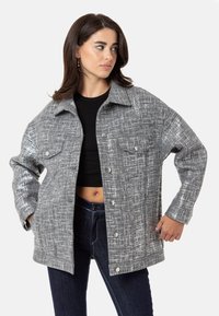 Grau strukturierte Jacke mit Kragen, Knopfverschluss und zwei Brusttaschen. Getragen über einem schwarzen Oberteil und kombiniert mit dunklen Jeans.