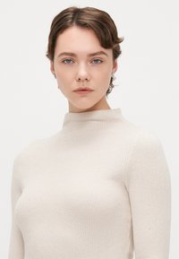 Pull à col montant et manches longues en tricot côtelé beige clair. Présente un design ajusté avec une texture subtile et une finition lisse.