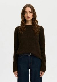 Donna con lunghi capelli castani mossi, che indossa un maglione fuzzy marrone scuro e jeans blu scuro, su uno sfondo chiaro e neutro.