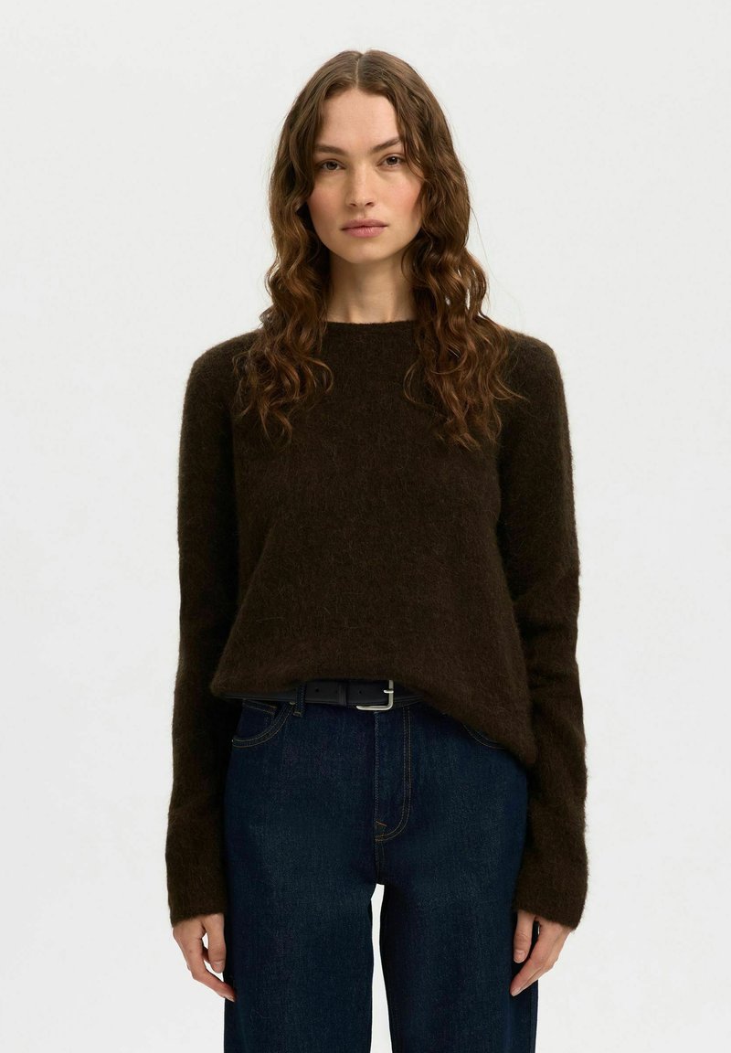 Donna con lunghi capelli castani mossi, che indossa un maglione fuzzy marrone scuro e jeans blu scuro, su uno sfondo chiaro e neutro.