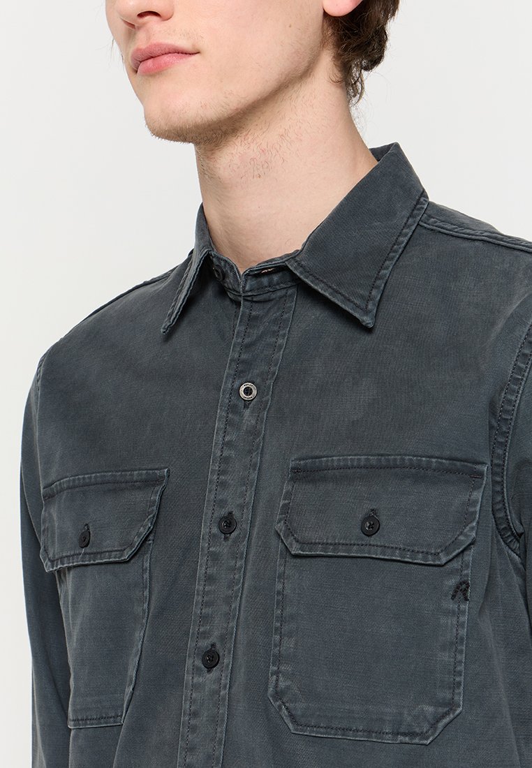 Junger Mann in einem dunkelgrauen Hemd mit Knopfleiste, zwei mit Knöpfen versehenen Vorderentaschen, Kragen und feiner Stoffstruktur.