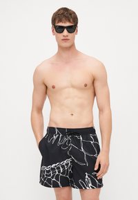 Sorte svømmebukser med et hvitt abstrakt design, som har et elastisk liv med snøring. Materialet virker lett og glatt.