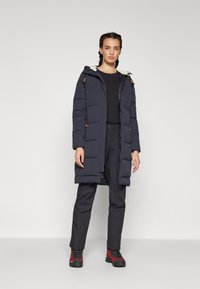 Cappotto imbottito blu con cappuccio, chiusura zip e dettagli arancioni. Indossato sopra una maglietta nera e pantaloni scuri, abbinato a calzature robuste con stivali rossi.