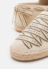 Espadrille en toile beige avec des rubans logo noirs. Présente un détail en jute tissé et un design à bout rond, mettant en valeur une tige en tissu texturé.