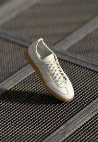 Sneaker bianca in pelle con tre strisce dorate, punta rotonda e suola in gomma beige. Presenta un design con lacci e il testo "Spezial" sul lato.