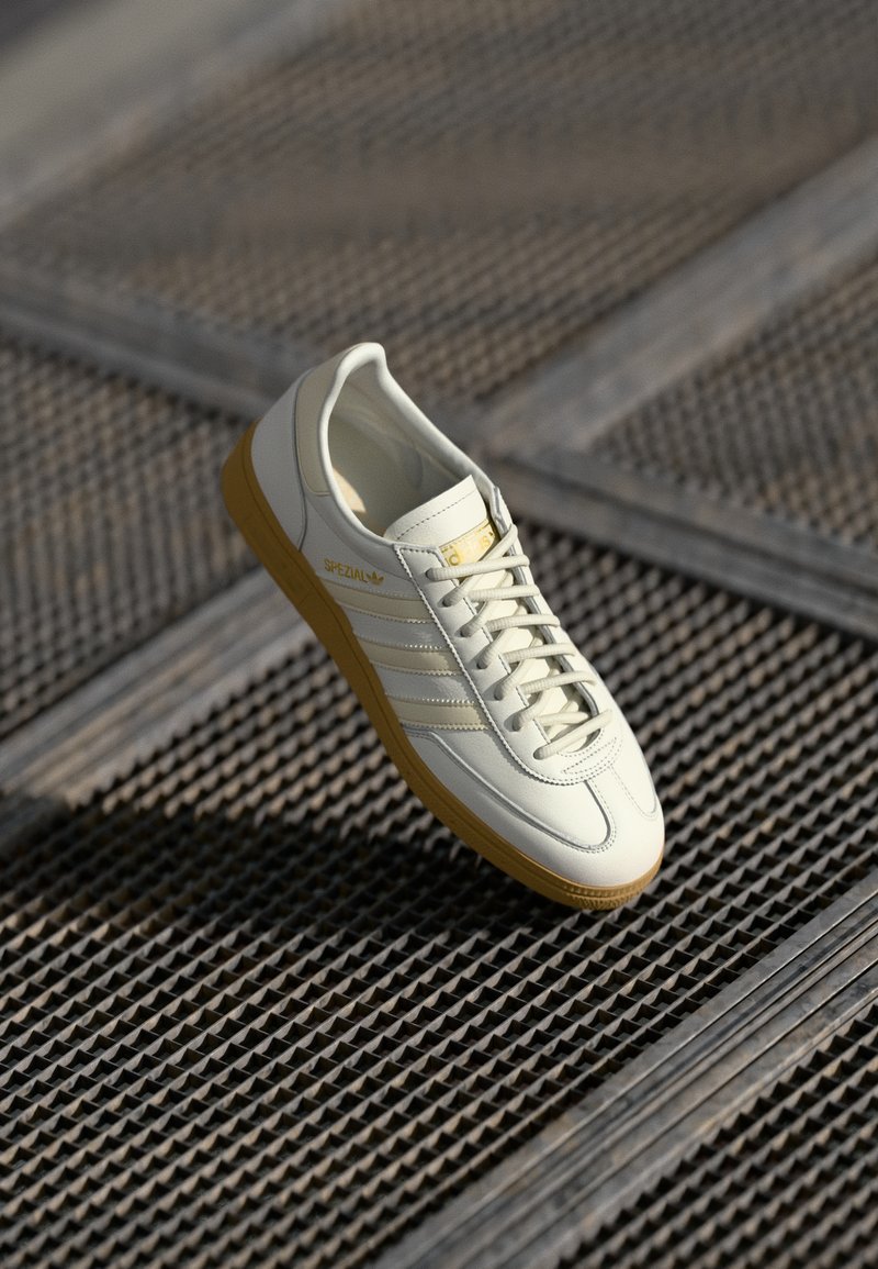 Sneaker bianca in pelle con tre strisce dorate, punta rotonda e suola in gomma beige. Presenta un design con lacci e il testo "Spezial" sul lato.
