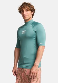 Τυρκουάζ κοντομάνικο rash guard με ψηλό γιακά, με λευκό λογότυπο στο στήθος. Λείο, ελαστικό ύφασμα για άνεση.