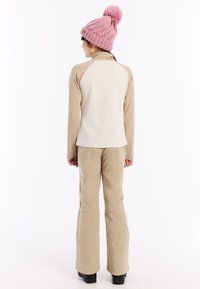 Beige skijack met roze accenten, gecombineerd met beige broek en zwarte laarzen. Een roze gebreide muts met een pompon maakt de outfit compleet.