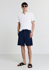 Uomo che indossa una maglietta bianca a maniche corte a costine, pantaloni corti navy con coulisse, sandali intrecciati neri e occhiali da sole quadrati scuri, in piedi con le mani nelle tasche.