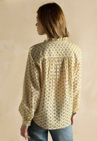 Bluza albă cu model floral galben, decolteu cu volane și detaliu adunat în spate. Mâneci lungi cu manșete, material ușor.