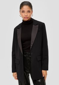 Black blazer with satin lapels, single button closure, two pockets, and a structured fit, paired with a black turtleneck and sequined trousers.