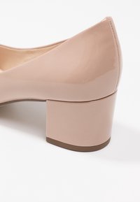 Nude lackläderpump med rundad tå och låg klack. Slät textur med en glänsande yta och subtila sömdetaljer.