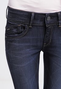 Jeans de ganga azul escura com um corte justo, apresentando uma cintura reta, bolsos frontais e costura amarela perto do bolso e da cintura.
