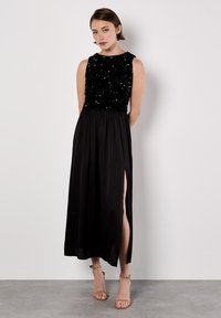 Abito da sera nero con un corpetto scintillante, design senza maniche e una gonna fluttuante con uno spacco laterale. Il modello indossa dei sandali nude con cinturini.