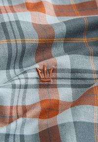 Sinise, halli ja oranži ruudulise mustriga riie, kootud tekstuuriga. Alumises osas on tikitud oranž kolmeharuline logo.