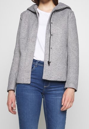 Femme portant une veste à capuche grise zippée par-dessus une chemise blanche et un jean bleu, debout devant un fond uni.