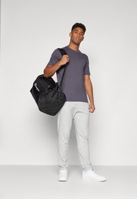 Sac de sport noir avec des accents en mesh et un logo, t-shirt gris, sweatpants gris clair et baskets blanches. Tenue décontractée de sport.