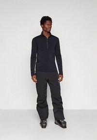 Férfi fekete hosszúujjú, félcipzáros felsőben, fekete hónadrágban és fekete síbakancsban áll egy világos háttér előtt.