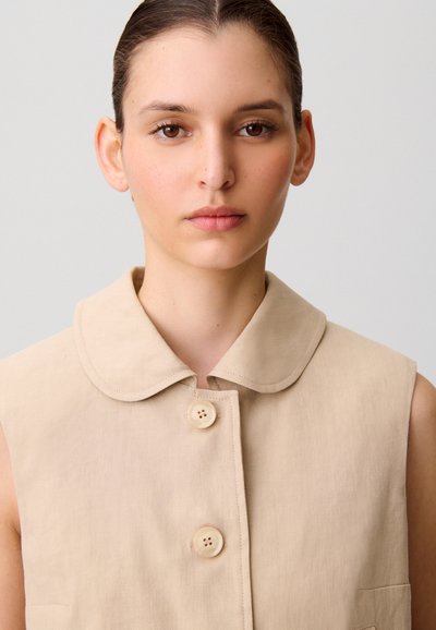 Giovane donna con occhi marroni e capelli raccolti, indossa un top beige senza maniche con bottoni e colletto arrotondato, sfondo neutro.