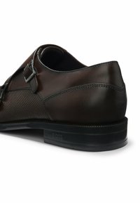 Dunkelbrauner Herrenschuh aus Leder mit Doppel-Monk-Riemen und geprägtem "BOSS"-Logo auf schwarzer Sohle, von der Rückseite gezeigt.