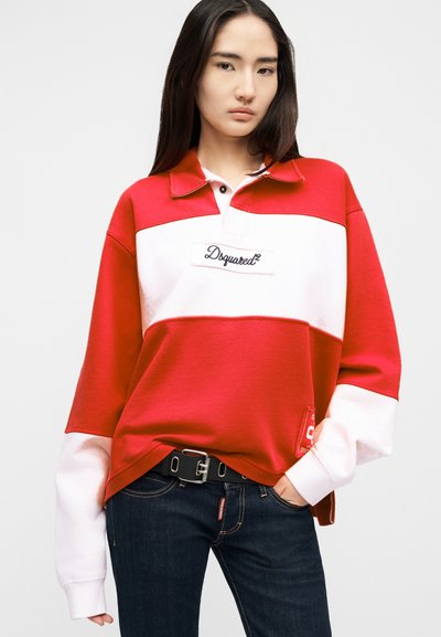 Γυναίκα με μακριά σκούρα μαλλιά, φορώντας κόκκινο και λευκό φούτερ Dsquared2 και σκούρο τζιν, στέκεται με το ένα χέρι στην τσέπη της.