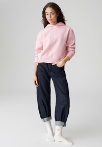 Lichtroze hoodie met een losse pasvorm, gecombineerd met donkerblauwe opgerolde jeans. Witte sokken met een klein roze hartje. Soepele stoftextuur.