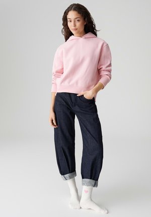 Hellrosa Kapuzenpullover mit lockerem Schnitt, kombiniert mit dunkelblauen, hochgekrempelten Jeans. Weiße Socken mit kleinem pinkfarbenen Herz. Glatte Stoffstruktur.