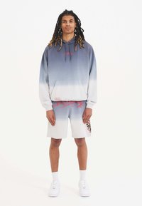 Sudadera con capucha de degradado azul, logo rojo; combinada con pantalones cortos gris claro con acentos florales y texto rojo en contraste. Zapatillas blancas.