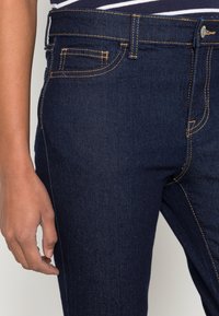 Los vaqueros de denim azul oscuro presentan un corte ajustado, un diseño de cinco bolsillos y costuras en amarillo contrastante a lo largo de las costuras y los bolsillos.