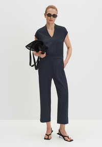 Donkerblauwe jumpsuit met kapmouwen en een V-hals. Strak model met een elastische tailleband. Gekoppeld met zwarte zonnebril en een netten tas.