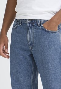 Siniset farkut, joissa on lievä haalistuma, klassinen viiden taskun muotoilu, vetoketjullinen haara ja nimetty nappi vyötäröllä.