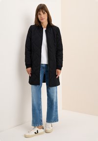 Zwarte gequiltte jas met een rits aan de voorkant, geribde mouwen en een knielange snit, gecombineerd met blauwe denim wijde jeans en witte sneakers.