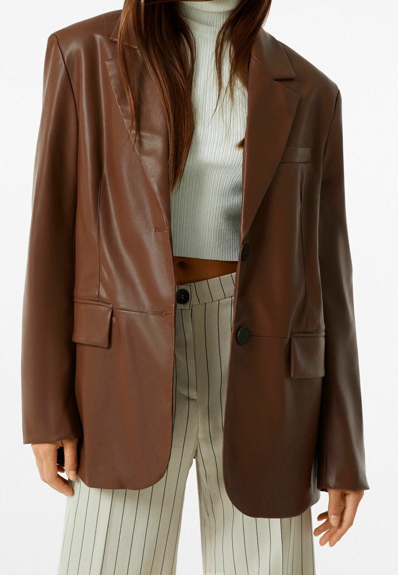 Brązowa marynarka z imitacji skóry o luźnym kroju, z klapami w szpic, dwoma przednimi kieszeniami i zapięciem na guziki, noszona na białym prążkowanym topie i spodniach w paski.