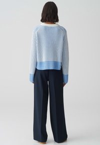 Person med mörkt hår som bär en blå och vit randig tröja och lösa svarta byxor, stående vänd bort från kameran mot en enfärgad bakgrund.