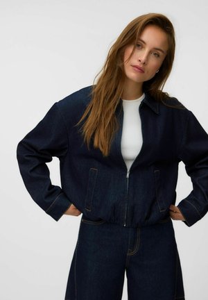 Femme aux longs cheveux bruns portant une veste en jean bleu foncé sur un haut blanc et un pantalon en jean bleu foncé assorti, debout, les mains sur les hanches.