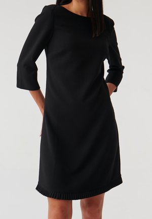 Czarna sukienka do kolan z rękawami do połowy przedramienia i plisowanym dołem. Wykonana z gładkiej tkaniny, o prostej, strukturalnej konstrukcji.