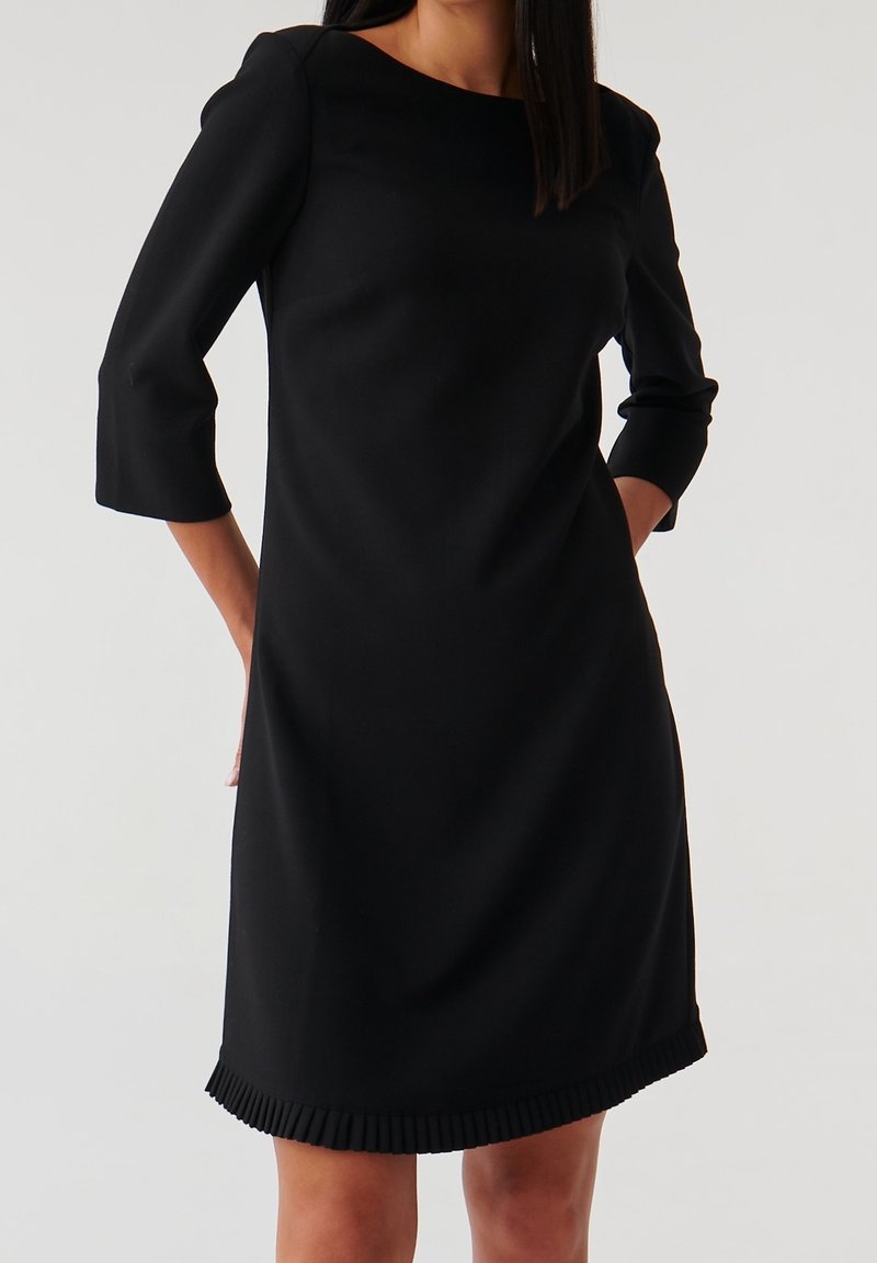 Robe noire longueur genoux avec manches trois-quarts et ourlet plissé. Confectionnée dans un tissu lisse, présentant un design simple et structuré.