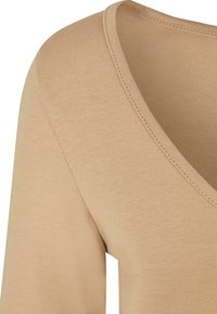 Beige Baumwolloberteil mit V-Ausschnitt und langen Ärmeln, ausgestattet mit einer gerippten Textur und sauberen Nähten an den Rändern.