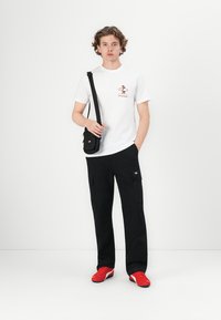 Hvid t-shirt med et brunt grafik, sorte cargo-bukser med lommer og røde sneakers. Lille sort crossbody taske. Enkel baggrund.