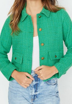 Frau trägt eine grüne, strukturierte Jacke mit Knöpfen über einem weißen Top und hellblauen, hoch taillierten Jeans, gezeigt von Hals bis Hüfte.