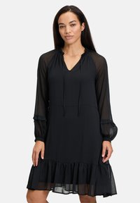Schwarzes Kleid mit transparenten langen Ärmeln, V-Ausschnitt, Kordelzug-Detail und einem rüschigen Saum. Weicher, leichter Stoff mit fließender Silhouette.