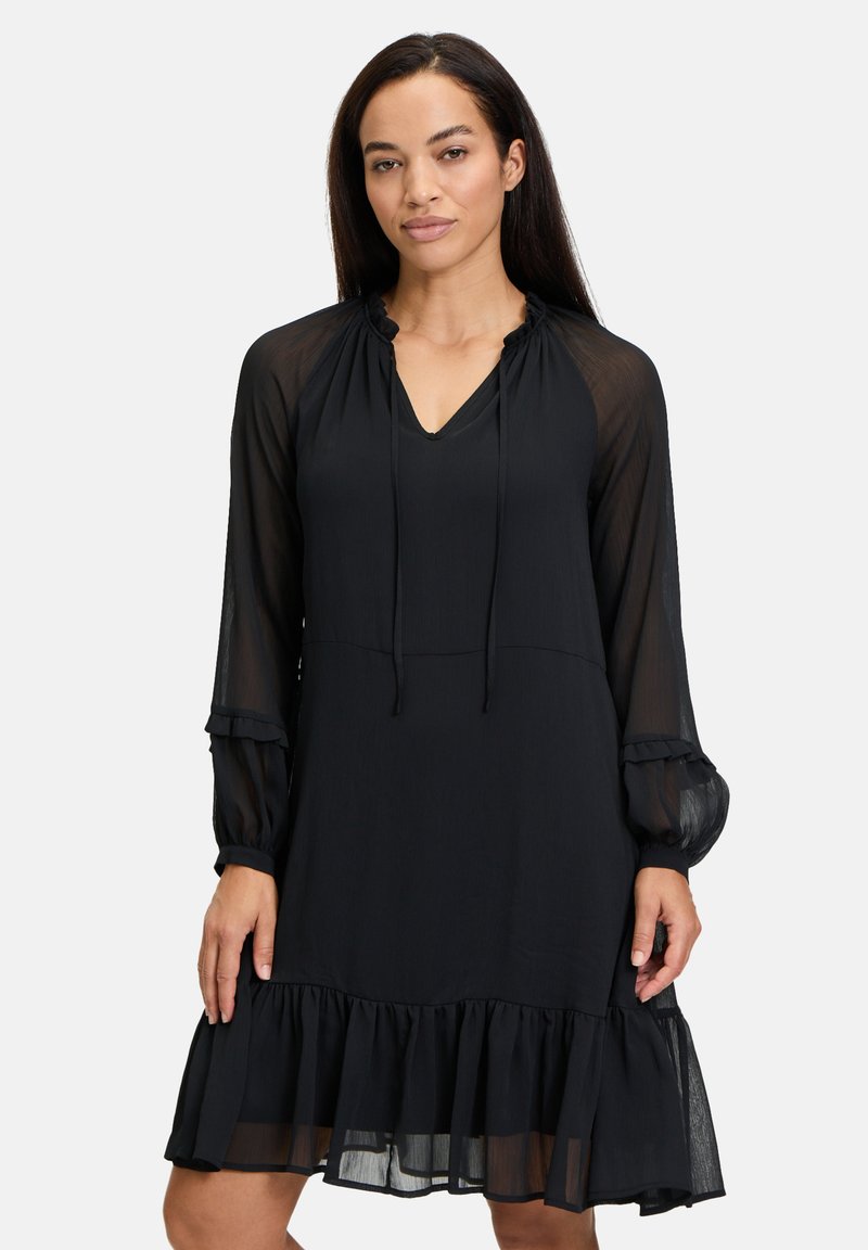 Schwarzes Kleid mit transparenten langen Ärmeln, V-Ausschnitt, Kordelzug-Detail und einem rüschigen Saum. Weicher, leichter Stoff mit fließender Silhouette.